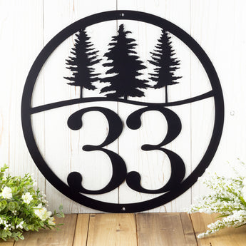 Close up of round metal house number sign with pine trees, in matte black powder coat.