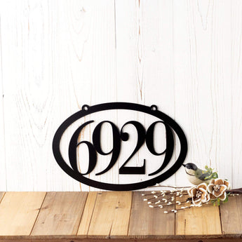 Horizontal oval 4 digit metal house number sign, in matte black powder coat. Placed against a white wood wall.