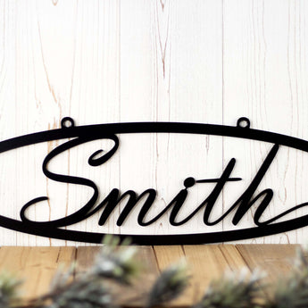 Close up of family last name on an oval metal sign with script font, in matte black powder coat. 