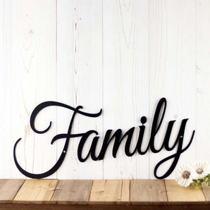 Family script metal sign, in matte black powder coat. Placed against a white wood wall.
