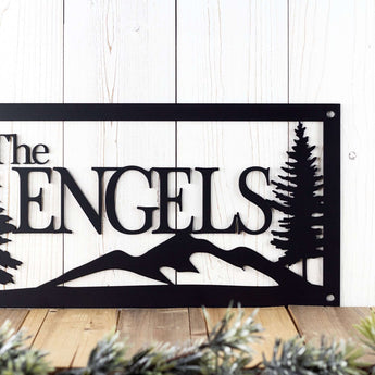 Close up of rectangular family name sign with pine trees and mountains, in matte black powder coat.