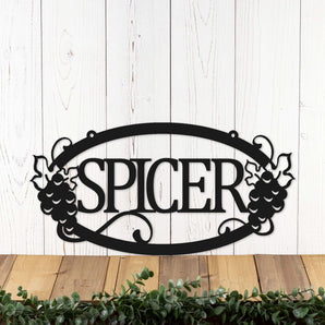 Hanging oval family name metal sign with grapes and grapevines, in matte black powder coat.