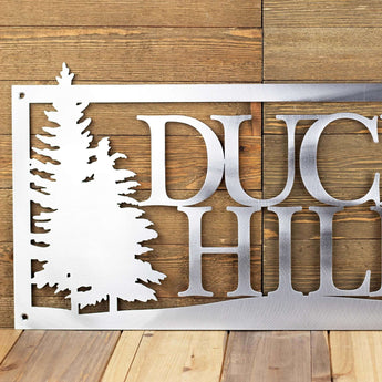 Close up of rectangular metal home name sign, with pine trees, in raw steel.