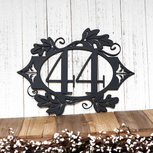 2 digit metal house number sign with oak leaves, in matte black powder coat. Placed against a white wood wall.